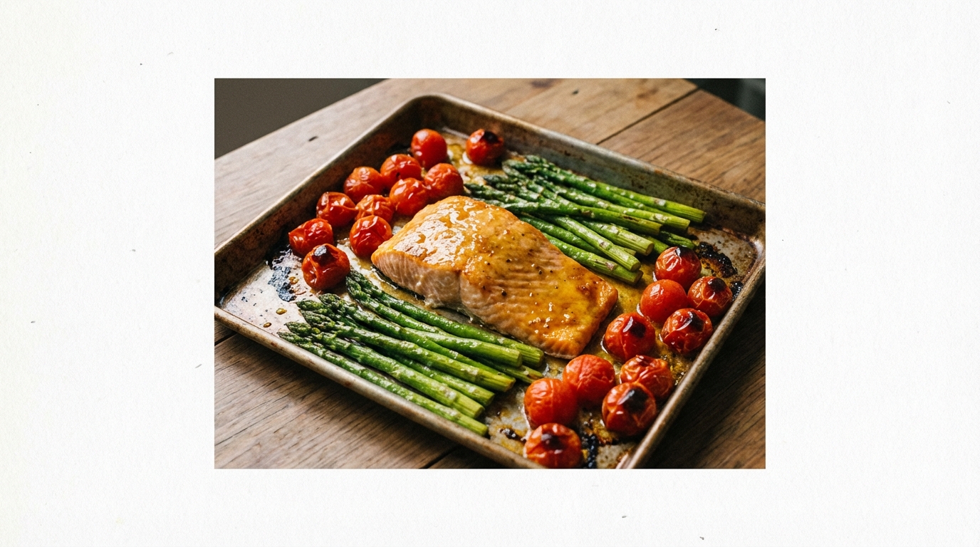 A beautifully roasted sheet pan dinner fresh out of the oven, featuring a side of salmon with a golden glaze, surrounded by roasted red cherry tomatoes and bright green asparagus.